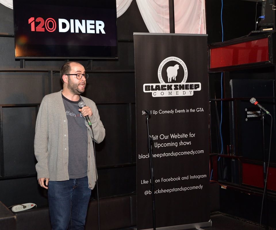 An image of Samuel Dunsiger on stage speaking into the microphone at 120 Diner.