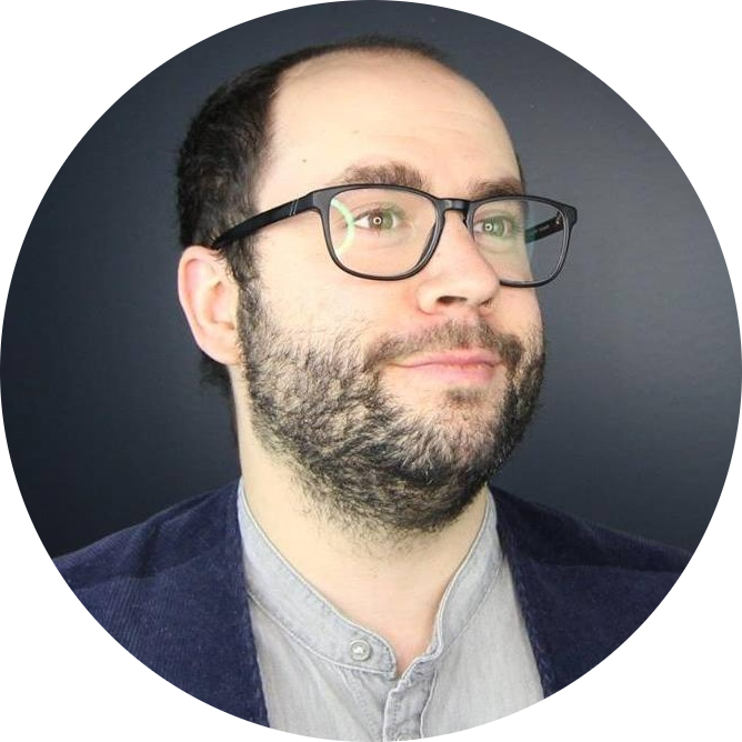 An image of Samuel Dunsiger, with a beard and glasses and wearing a grey shirt and vest. He is seen looking out and smiling with a dark grey background.